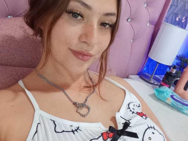 SamanthaCollen with freckles, wearing a white Hello Kitty top, silver chain necklace with heart pendant, brown hair, and subtle pink lipstick, smiling at the camera.