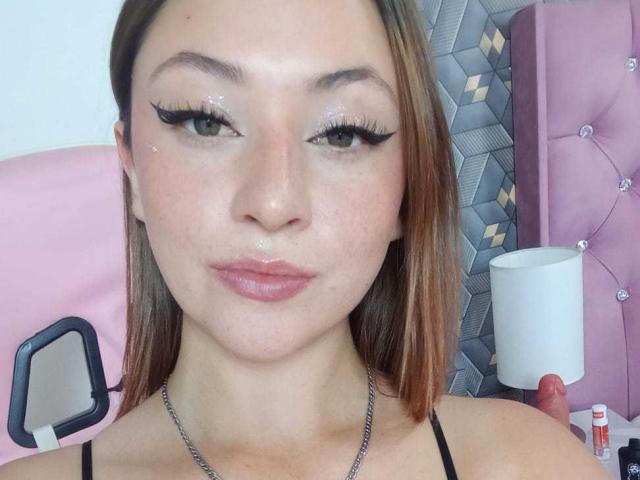 SamanthaCollen has shiny eyelids, thick black eyeliner, and glossy pink lips. She's wearing a black strap top, with a chain necklace, and has a small droplet on her nose.