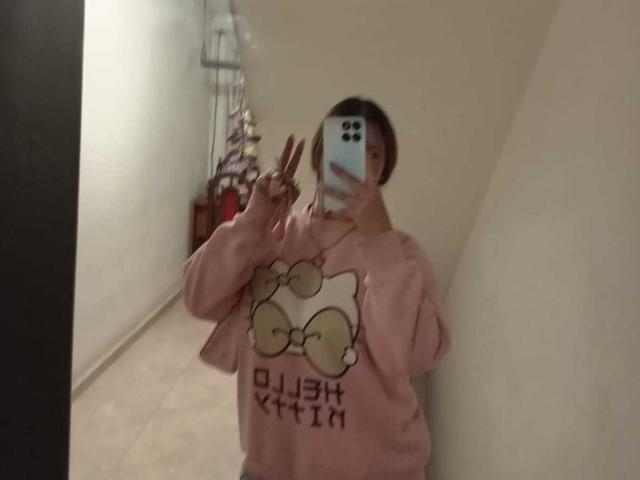 SamanthaCollen in a pink Hello Kitty sweatshirt, making a peace sign with her right hand, holding a phone in front of her face, in a dimly lit room.