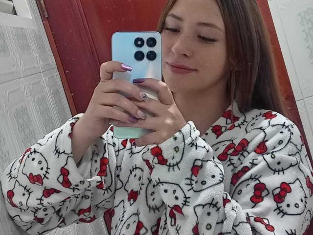 SamanthaCollen in a Hello Kitty robe, taking a selfie with a blue iPhone, eyes closed, slight smile, long brown hair, purple nails, holding phone with three cameras.