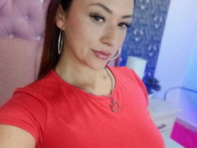 SamanthaCollen with brown hair, wearing a red shirt, hoop earrings, and a necklace with a heart pendant, showing her tits, smiling with glossy pink lips.