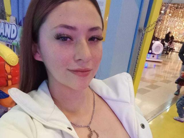SamanthaCollen with long brown hair, white jacket, and chain necklace, showing off her small tits in a selfie. She's smiling with pink eyeshadow and long lashes.