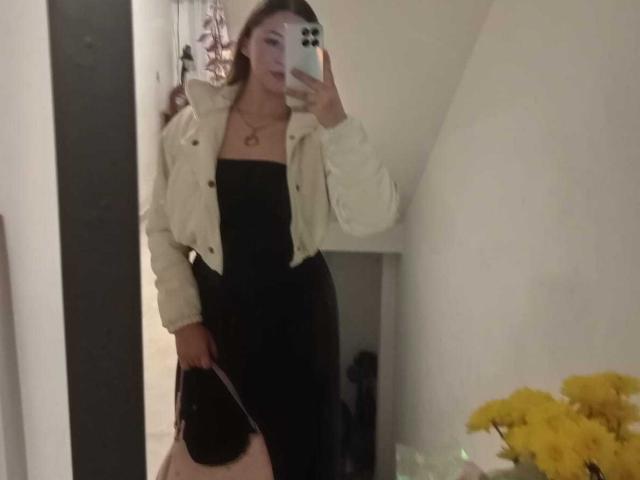 SamanthaCollen with long brown hair, wearing a black strapless dress and beige jacket, holding a black purse, taking a mirror selfie, showing her tits and ass.