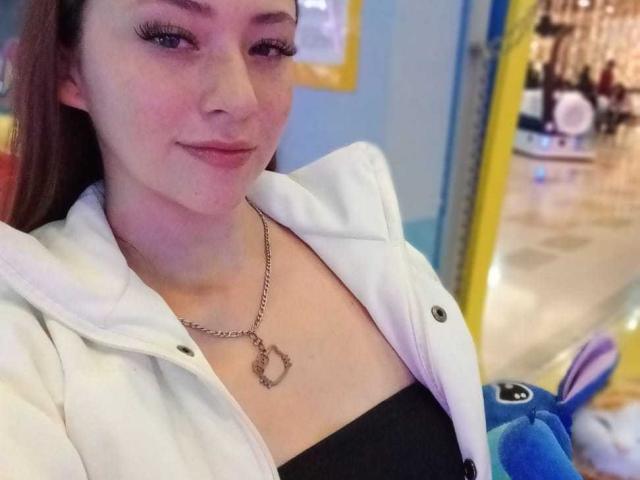 SamanthaCollen with fair skin, long brown hair, and a small smile, wearing a white jacket over a black top, a chain necklace with a heart-shaped pendant, and blue plush toy.