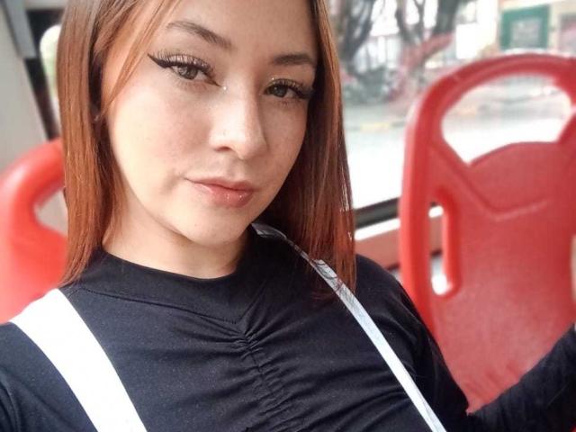 SamanthaCollen with medium brown hair, black top, white straps, eyeliner, and a slight smile, holding a straw, red plastic chair in the background.
