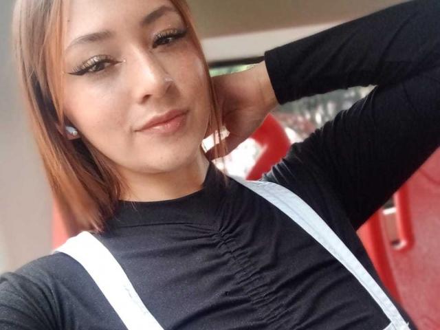 SamanthaCollen with light brown hair, black long-sleeve shirt, white suspenders, eyeliner, and earrings, hand behind head, smiling slightly, visible nose ring, small breasts.