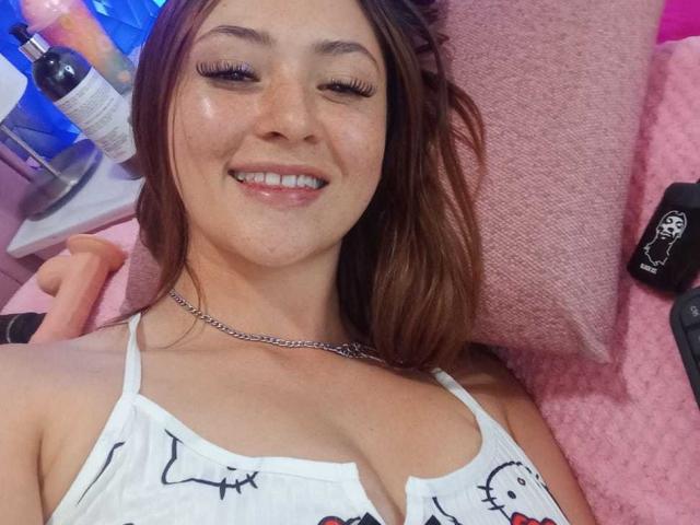 SamanthaCollen with wavy brown hair, white tank top, and chain necklace, smiling at camera, winking, showing off her shiny freckled face and small to medium tits.