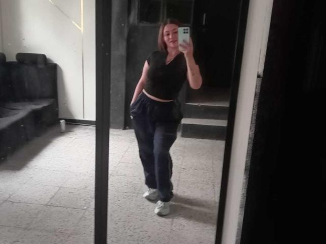 SamanthaCollen in a mirror selfie, wearing a black crop top, loose dark pants, and gray sneakers, with her hand in her pocket, standing with a slightly awkward posture.