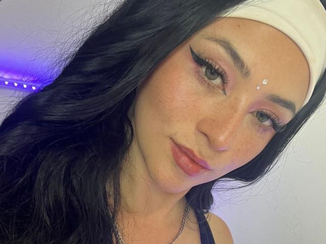 SamanthaCollen with long black hair, white headscarf, eyeliner, freckles, and small facial gems, looking at camera with pouty lips, wearing a black top.