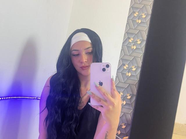 SamanthaCollen with long black hair, white headband, black tank top, holding iPhone, taking mirror selfie, looking down, light purple and blue lights, hexagon mirror on right.