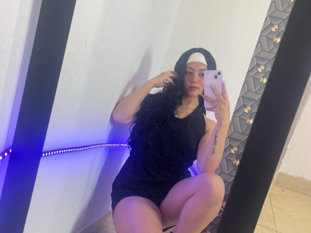 SamanthaCollen with long black hair, wearing a black tank top and beanie, squatting in front of a mirror, holding a phone, showing her ass and a tattoo on her arm.