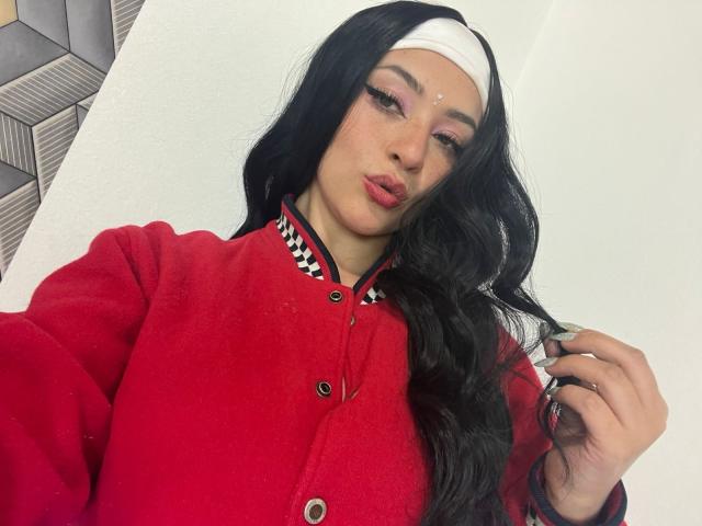 SamanthaCollen with long black hair, red button-up jacket, white headband, pink eyeshadow, and red lipstick, holding hair, looking at camera, white wall in background.