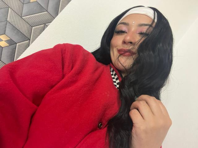 SamanthaCollen with long black hair, wearing a red cardigan and white headband, pouting with pink lipstick, holding hair, looking at camera, face adorned with small white dot.