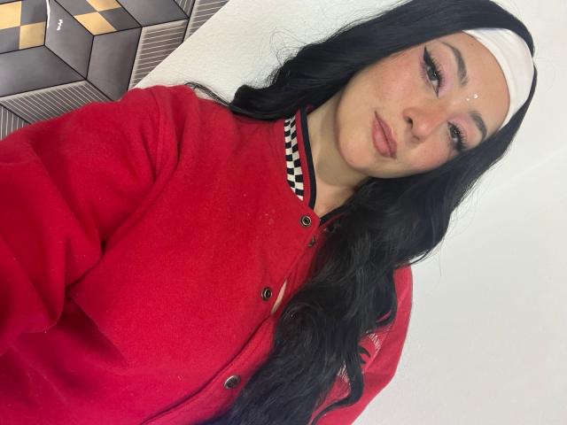 SamanthaCollen with long black hair, white headband, red cardigan, checkered collar, subtle makeup, and a small white mark on her forehead, looking at the camera.