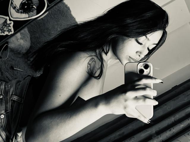 Schwarz-weißes Selfie von KatianPierce mit langen, dunklen Haaren, die ein Oberteil trägt, das ihre kleinen Brüste zeigt, und ein Telefon hält. Sie hat ein Tattoo an der rechten Schulter, trägt Jeans und dunkle Nägel..