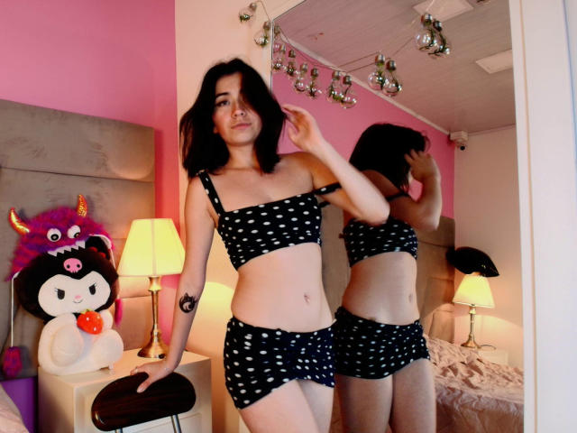 AnyuWu, con i capelli scuri, indossa un reggiseno e una gonna a pois neri, ed è in piedi di fronte a uno specchio che riflette il suo sedere e i suoi seni. La stanza è rosa, c'è una lampada e un peluche..