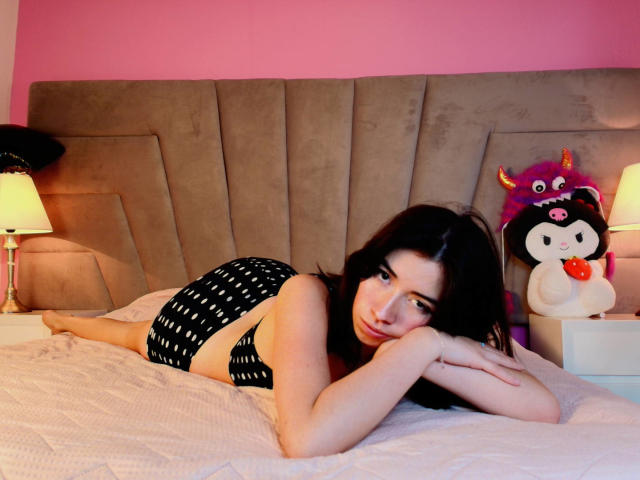 AnyuWu está acostada en una cama, vestida con bombachas negras con lunares, tiene los pechos pequeños, cabello negro, y está mirando a la cámara, en una habitación con paredes rosas, al lado de un juguete monstruo morado..
