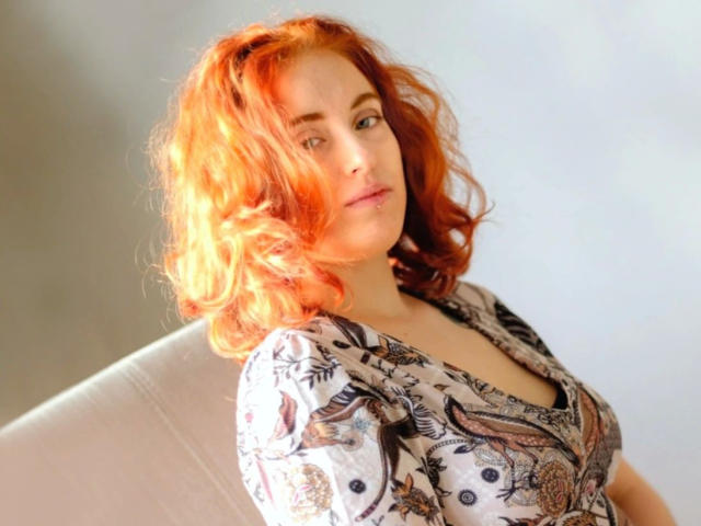 RubyLust-hot with bright orange wavy hair, wearing a floral-patterned top, looking slightly to the side with a soft expression. She has fair skin and small breasts.