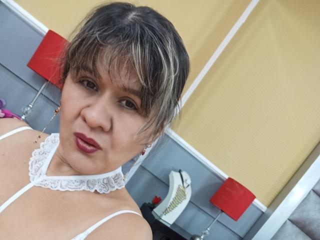 SophySheikh with short, blonde-streaked hair, wearing a white lace choker and red lipstick, looking at the camera, showing her tits, in a yellow and gray room.