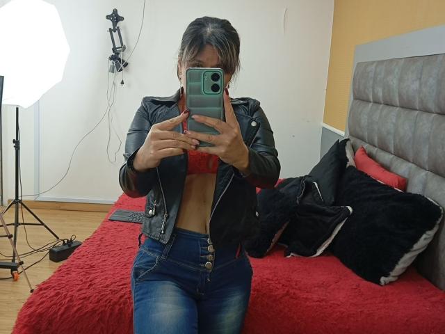 SophySheikh in a black leather jacket, blue jeans, and red bra, sitting on a red bed, taking a mirror selfie with a green phone, showing her tits.