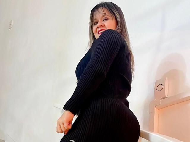 SophySheikh with long brown hair, wearing a black ribbed dress, showing her curvy ass and smiling at the camera. She's sitting against a white wall.
