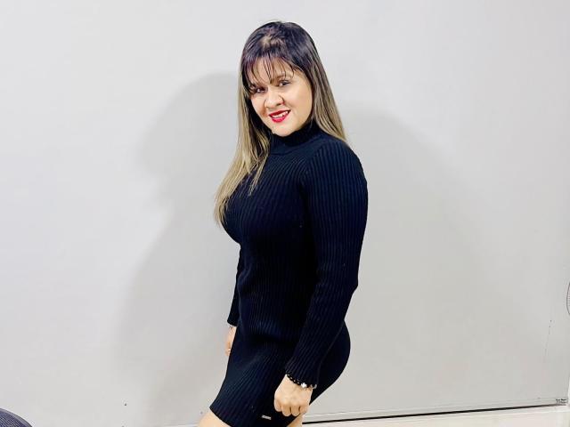 SophySheikh has long brown hair, red lipstick, and a black rib-knit dress, showing her curvy ass and small tits, smiling at the camera.