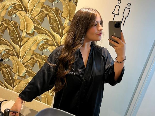 LorensDouce with long brown hair, wearing a black salon robe, taking a mirror selfie with a smartphone, showing a cute face and a bracelet on her wrist.