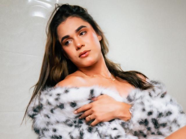 AliceCoolenk is a young woman with long, wavy brown hair and subtle makeup. She has a light tan complexion and is looking slightly to the left with a neutral expression. She wears a fluffy, off-the-shoulder, black-and-white leopard print coat, which she holds over her chest with her right hand. Her nails are manicured and she has a small gold necklace. The image is softly focused, with a slight blur on the edges, giving it a dreamy, almost ethereal quality. The lighting is even, highlighting her facial features and the texture of the coat.