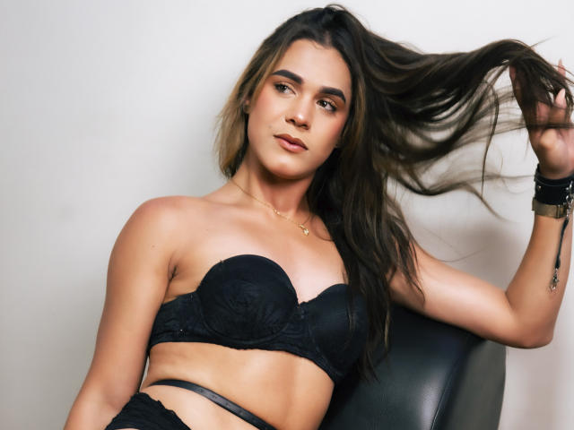 AliceCoolenk, a young woman with long brown hair, holds her hair in her right hand. She wears a black strapless lace bra and a black skirt, revealing a slim, toned midsection. She has a light olive skin tone, dark eyebrows, and subtle makeup. A small gold necklace and a black wristband with a gold buckle are visible. Her expression is calm, and her gaze is directed at the camera. The image is sharp with a neutral, slightly off-white background. The composition focuses on her upper body.