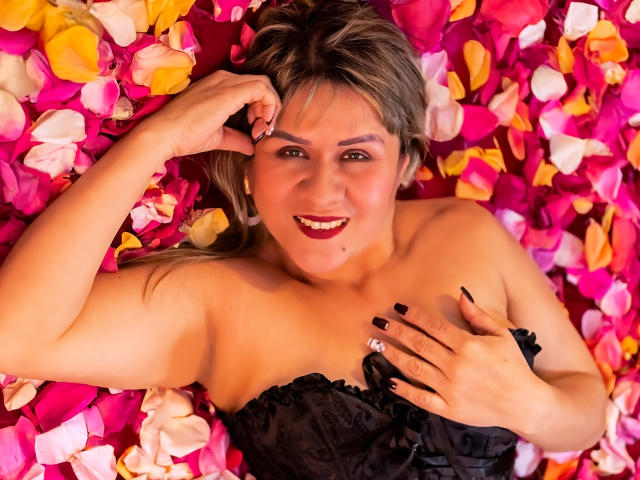 SophySheikh lying on a bed of pink and yellow petals, smiling with dark lipstick, black strapless dress, black nails, hand touching her chest, blonde hair, fair skin.
