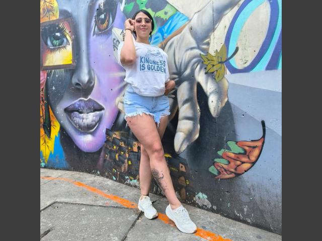ZaraBeicker with her ass up, wearing a white shirt and blue denim shorts, has a tattoo on her left thigh, and white sneakers, standing in front of a graffiti wall.