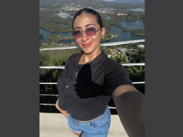 ZaraBeicker with tan skin, black button-up shirt, blue shorts, brown sunglasses, smiling, hands on hips, standing outdoors, greenery and water in background, selfie, small shadow on her shirt.
