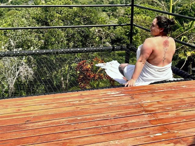 EmiliaMora with her back to the camera, showing a tattoo down her spine, sits on a wooden deck, wrapped in a white towel, exposing her bare ass.