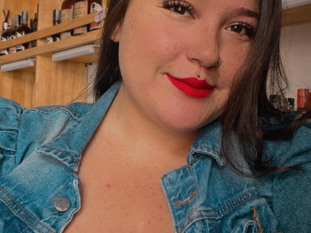 EmiliaMora with red lips, nose ring, and denim jacket, showing her chest, has brown hair, and is smiling slightly. Her tits are visible under the jacket.