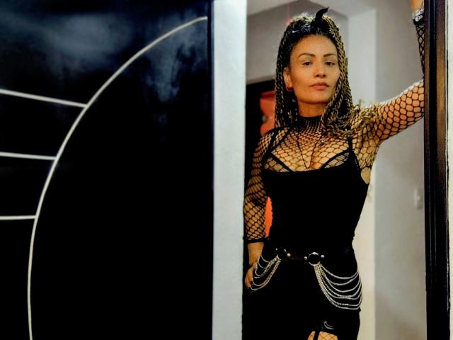 DivaMarie with braided hair, wearing a black fishnet top, showing her tits, and a black dress with chain belts, standing in a doorway, looking confident.