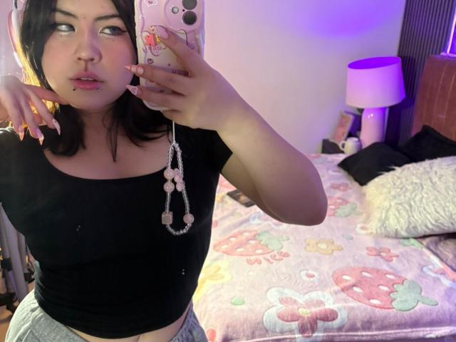 HoneeyBonny with dark hair, black crop top, and pink nail polish, showing her face and nipple piercing, holding a phone with a pink case in a bedroom selfie.