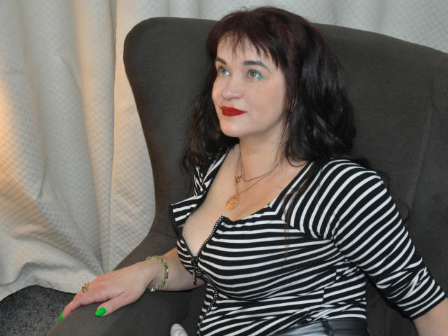 CharlotteFlori sits in a dark gray armchair with white curtains in the background. She has wavy, dark brown hair and bright red lipstick. She wears a black and white striped, low-cut top that reveals a hint of cleavage, and a gold necklace with a small pendant. Her green-painted nails rest on the armchair. The image has a casual, everyday feel. Her light skin and blue eyes are prominent. She looks slightly to the right with a subtle smile. The overall composition is simple and focused on CharlotteFlori.
