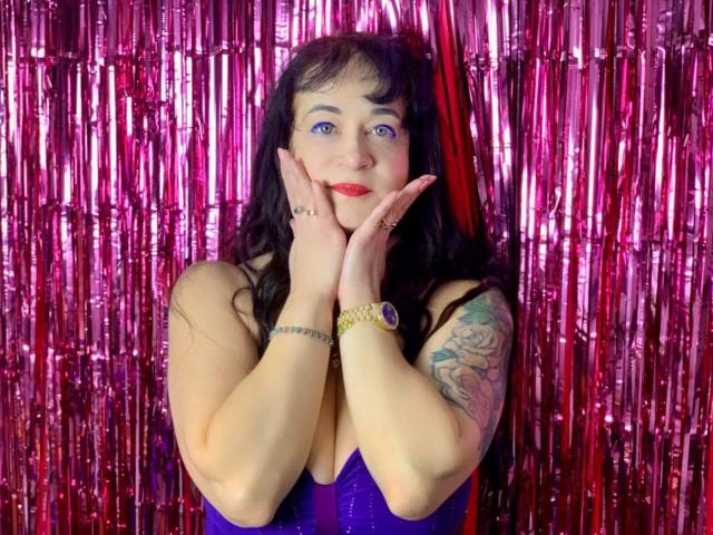 CharlotteFlori with purple dress, red lipstick, black hair, blue eyes, hands up, tattoo on right arm, pink and red metallic curtain, watch on left wrist, smiling.