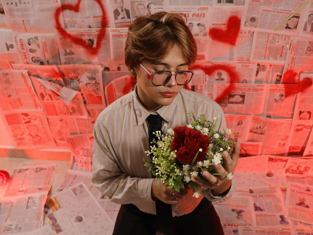 BenjiVampire with short brown hair, glasses, striped shirt, and tie, holding a red rose bouquet with greenery, red hearts in the background, and red lighting.