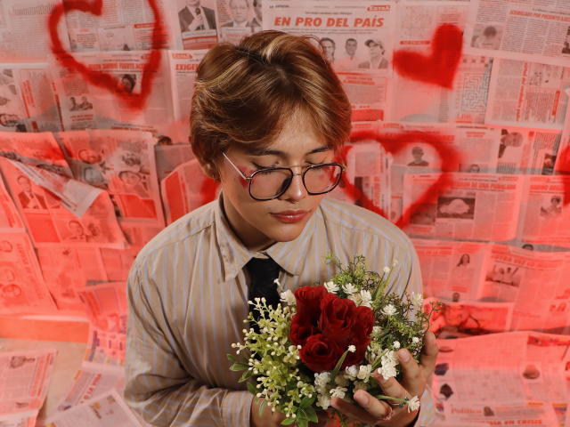 BenjiVampire with short brown hair, glasses, striped shirt, and black tie, holding a bouquet of red roses and white flowers, looking down. Red heart graffiti in background.