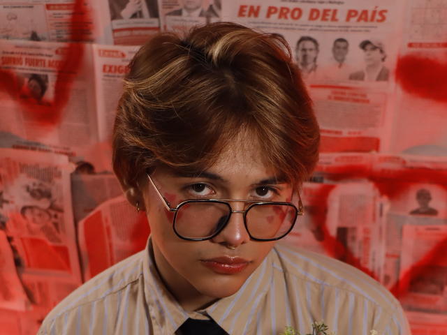 BenjiVampire has short brown hair, wears glasses, a striped shirt, and a nose ring. Red graffiti is on the wall behind. She has red lipstick and a serious expression.