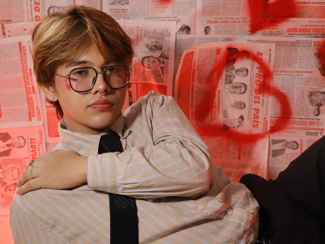 BenjiVampire with glasses, striped shirt, black tie, arms crossed, looking at camera, red graffiti on background, brown hair, subtle smile, standing, ring visible on right hand.