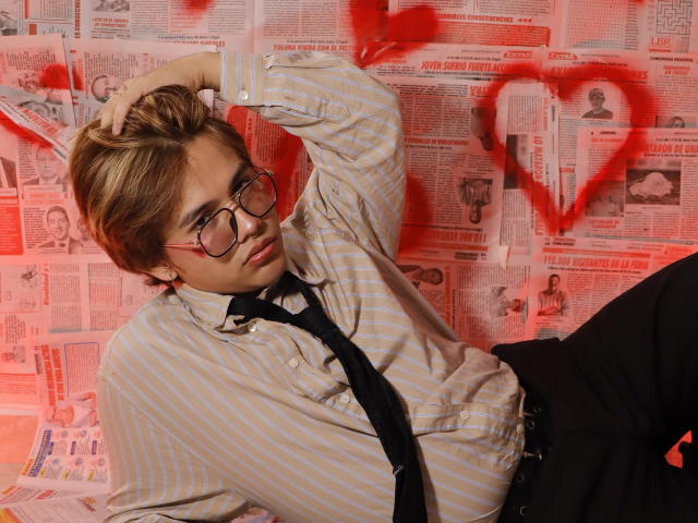 BenjiVampire with glasses, striped shirt, black tie, and loose black pants, hand in hair, red heart graffiti, looking at camera, sexy, nerdy, slightly messy, casual pose.