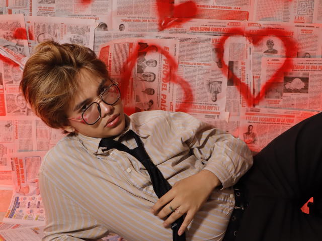 BenjiVampire with short blonde hair, glasses, striped shirt, black tie, and black pants, leaning against newspaper wall with red heart graffiti, looking at camera, ring on left hand.