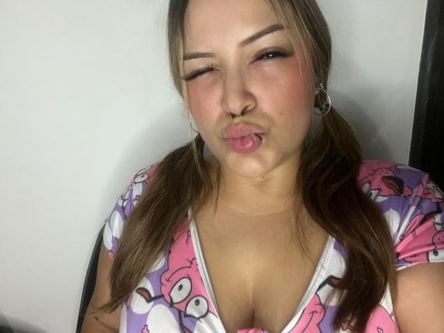 AllisonJustingb tiene labios rosados y voluminosos, un piercing en la nariz y coletas marrones. Lleva una blusa con un estampado de dibujos animados en rosa y morado, que deja ver sus grandes senos. Está guiñando un ojo y sacando la lengua..