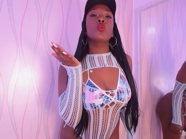 BriannaThompson with big ass, wearing a white, fishnet, cut-out top showing her tits, black cap, long nails, and hoop earrings, pouting with pink lips, in a pink-lit room.