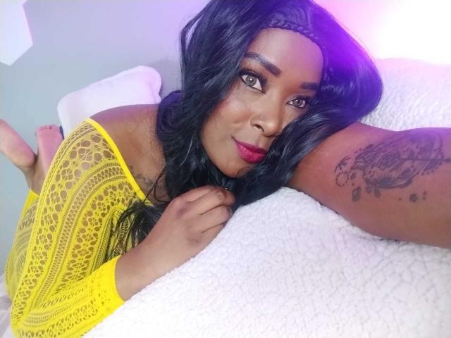 BriannaThompson lying on a white bed, wearing a yellow lace top, exposing her shoulder tattoo, with her black hair and red lipstick. Her pussy is not visible.