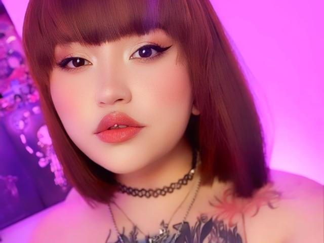 AliBrinck with shiny brown bob, pink eye makeup, glossy red lips, black choker, tattoos on chest, pink and purple lighting, and a sexy, cute expression.