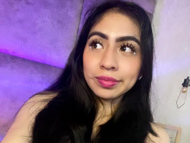 ZoeRedds with long black hair, brown eyes, and pink lipstick, looking to the side with a slight smile. She has a small beauty mark under her right eye.