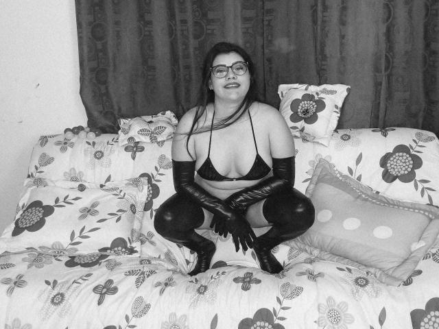 ValeriaLoveer, kurvig mit großen Brüsten, sitzt auf einem Blumenbeet in einem schwarzen BH, Handschuhen und Stiefeln, lächelnd, mit Brille, langem, dunklem Haar, auf einem Bett mit gemusterten Kissen..